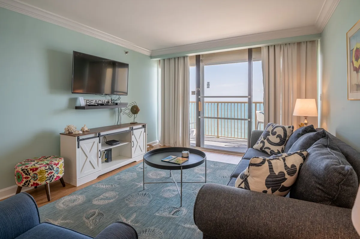 A bright, cheerful living room featuring a plush sofa and coffee table, with a large TV and glass doors leading out to a balcony. Sunlight streams in, and the inviting atmosphere is perfect for relaxing, watching movies, or chatting with friends and family. The balcony and ocean view are visible, tying inside and outside together.