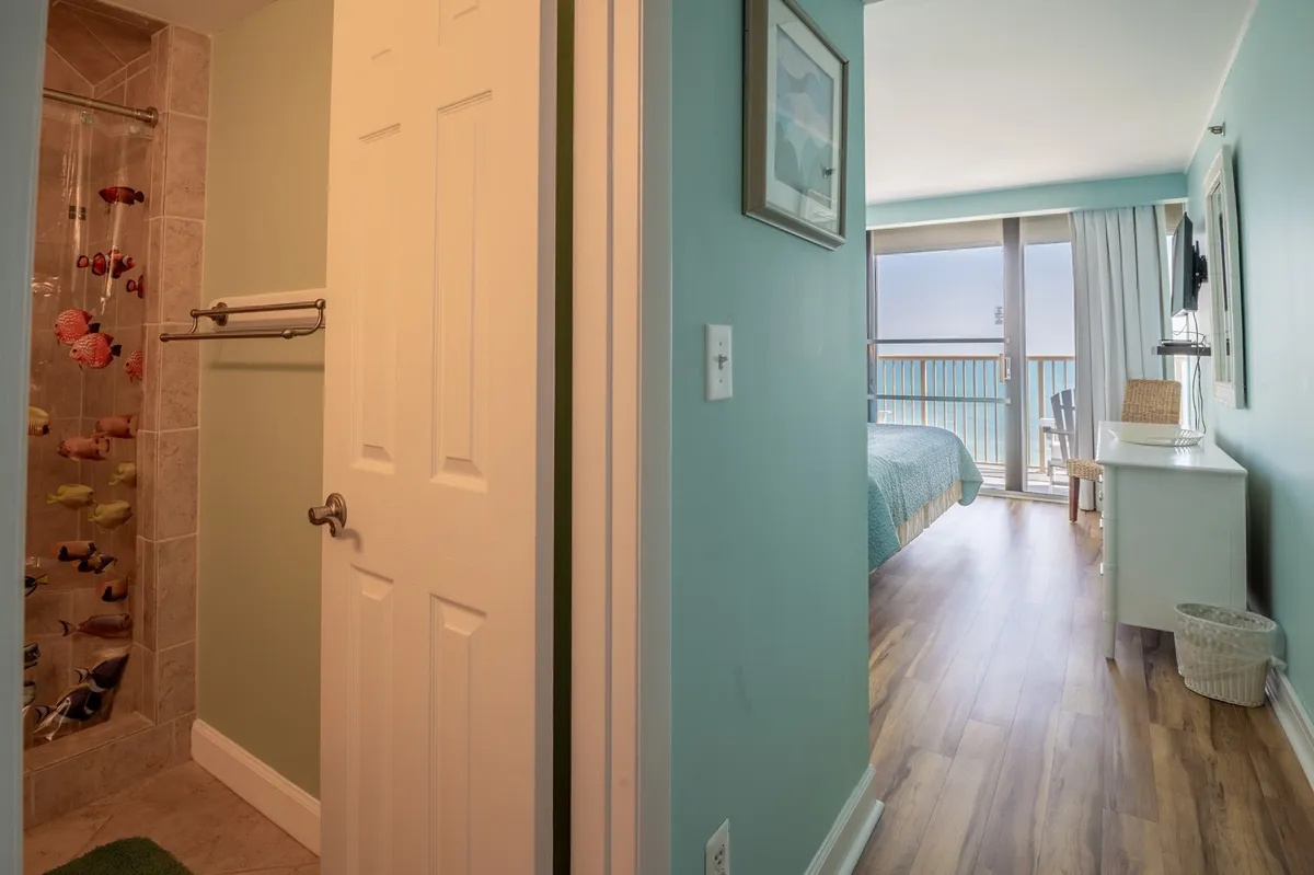A bright bedroom with a view of the balcony, colorful wall art, and a shower area with vibrant accessories. The bedroom opens up to outdoor views, creating a sense of connection to the beach environment.