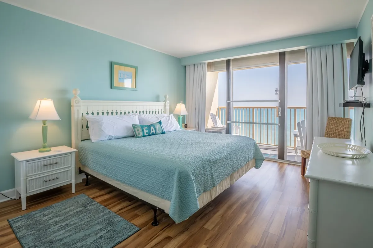 A serene bedroom featuring a king-sized bed, decorative pillows, a nightstand with a lamp, and floor-to-ceiling glass doors opening out to a private balcony with sweeping sea views. The soft lighting and tranquil decor evoke a peaceful retreat ideal for unwinding after a day of adventure.