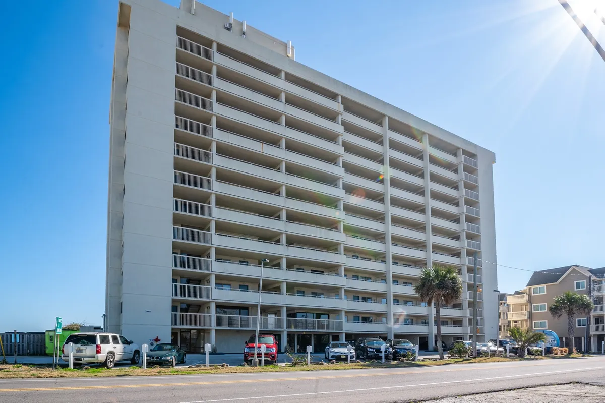 This image features the exterior of a tall building with open balconies and cars parked in front. The building's prominent entrance and visible parking reinforce the ease of access for arrivals and departures.