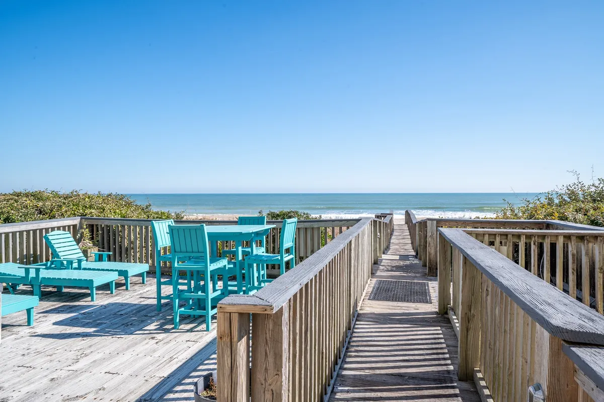 A wooden deck furnished with vibrant turquoise chairs overlooks the ocean, with ample space for sunbathing or relaxing. The scene is bright, with sunlight enhancing the cheerful mood. The open view connects directly to the beach, inviting guests to enjoy both daytime lounging and sunset gatherings.