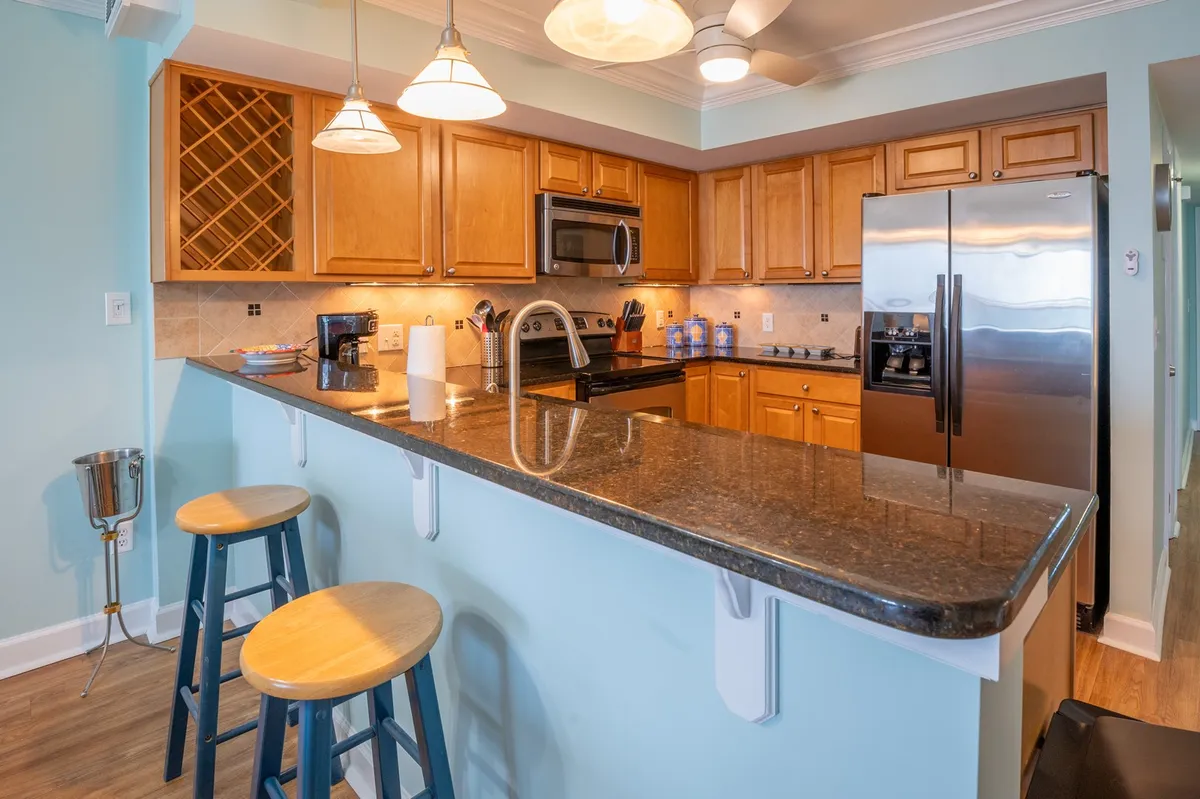 A warm, modern kitchen with granite countertops, stainless steel appliances, and a sleek breakfast bar with bar stools. The space is ready for cooking or casual breakfasts, keeping the chef in the center of social activity.