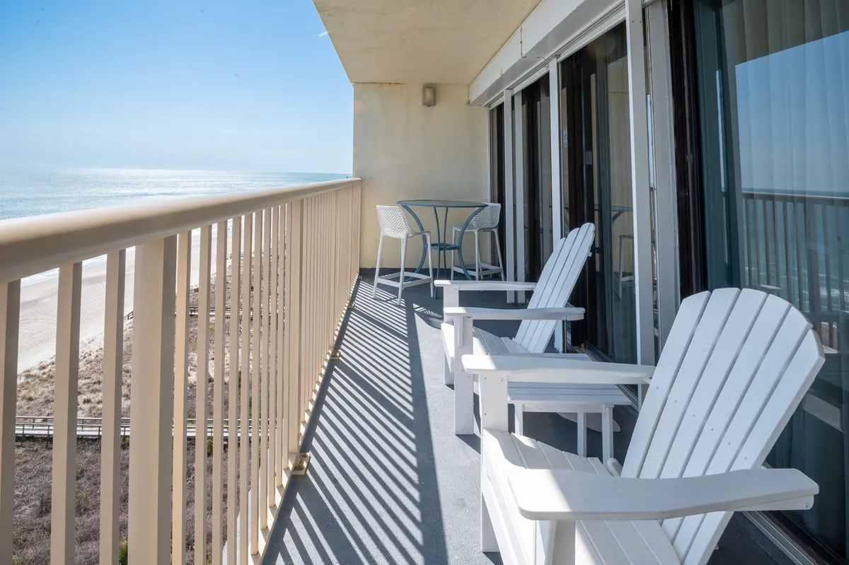 White Adirondack chairs and a small table are set on a private balcony facing the ocean. The tranquil setting is ideal for sipping drinks, enjoying the breeze, or watching the sunrise and sunset, evoking a sense of private retreat before or after exploring local nightlife.