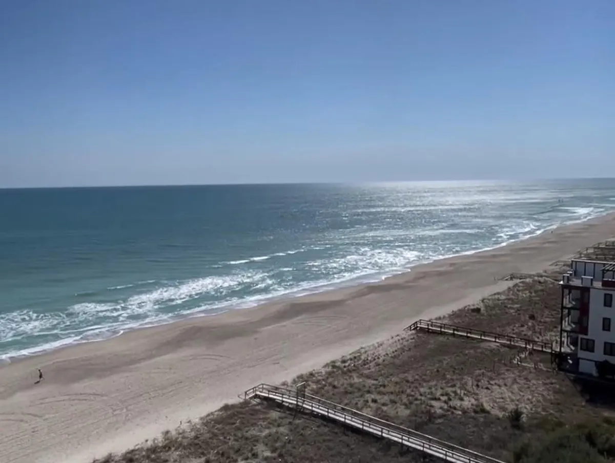 A sweeping view of the sandy beach, rolling ocean waves, a distant boardwalk, and the wide horizon. The scene is calm and open, inviting families to imagine themselves on the sand, engaging in activities like beachcombing, sunbathing, or playing near the water.
