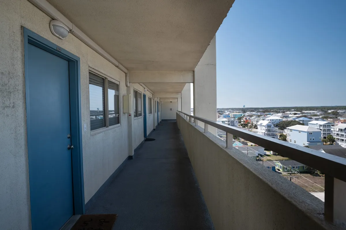 This image shows a blue door, balcony railing, and surrounding buildings, capturing the exterior corridor leading to the entrance. The scene hints at the transition from the parking area up to the unit, highlighting accessibility and proximity to amenities.