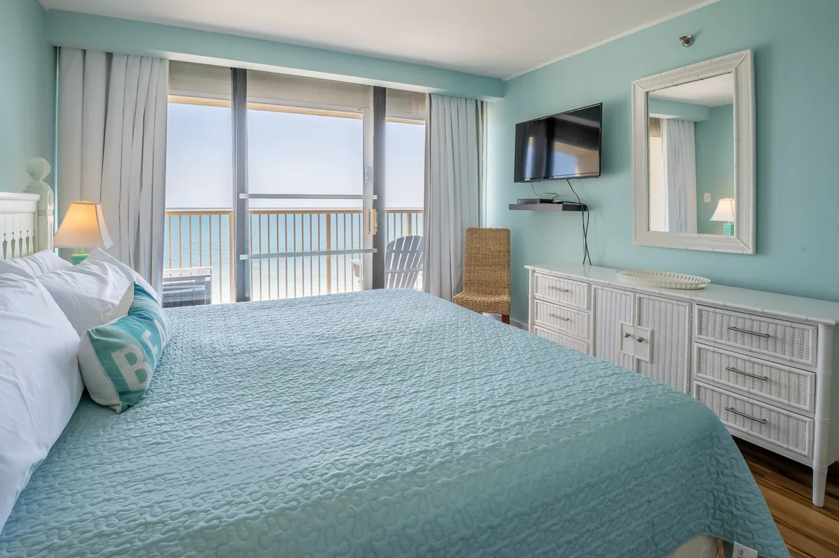 A bright, inviting bedroom with a queen-sized bed covered in a turquoise blanket, large windows offering panoramic ocean views, a mounted TV, a white dresser, and a cozy wicker chair. Natural light fills the space, creating a welcoming environment.
