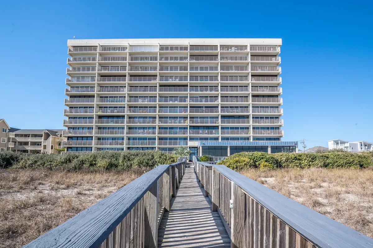 This exterior image features a wooden walkway leading from the modern building toward the beach, surrounded by lush greenery. The view captures the direct boardwalk access guests enjoy from the property, setting a tranquil and inviting scene for families preparing for a day by the ocean.