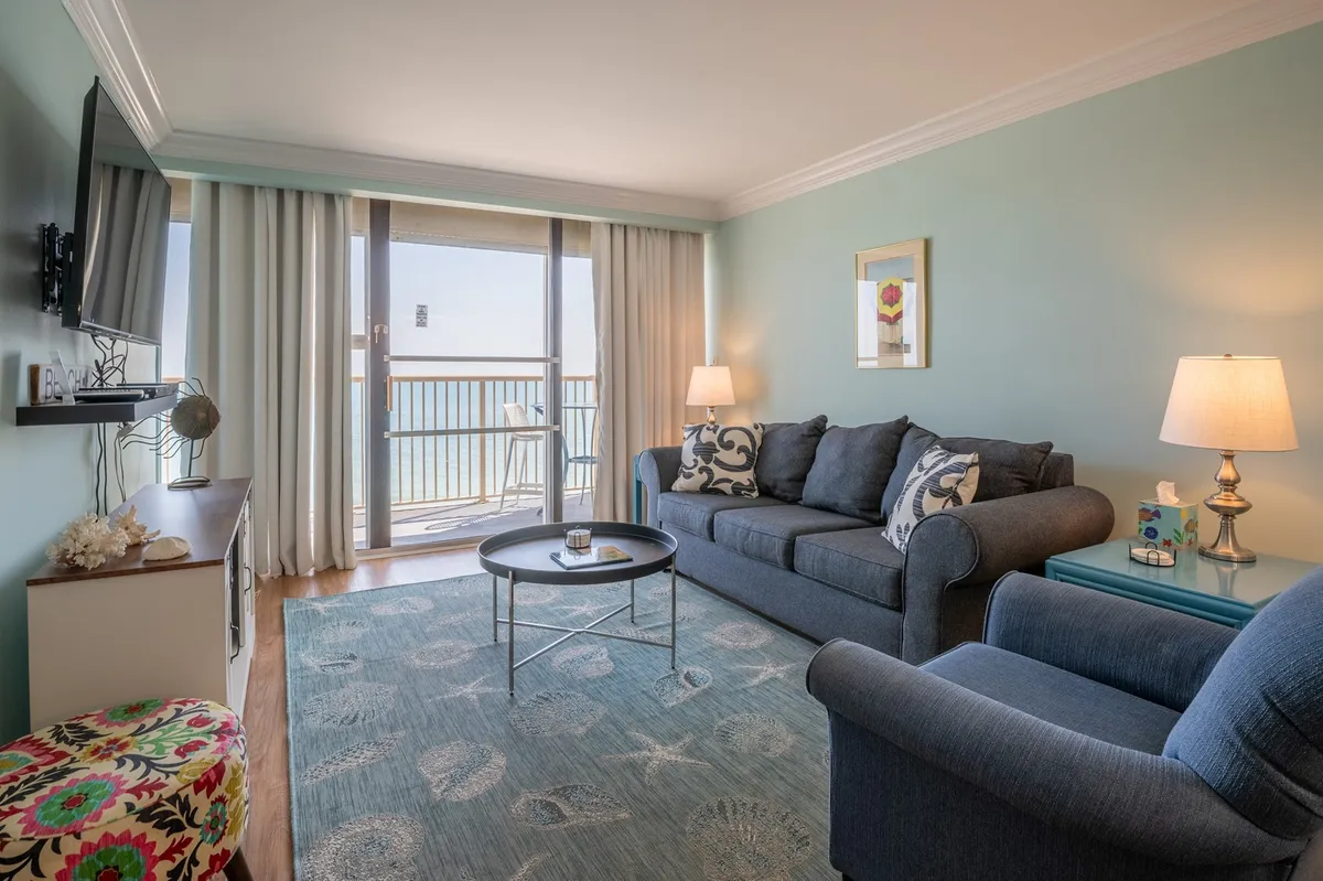 A cozy interior living room with a sofa, coffee table, decorative lamp, and a view out onto the balcony. The scene suggests a seamless blend between indoor comfort and outdoor vistas.