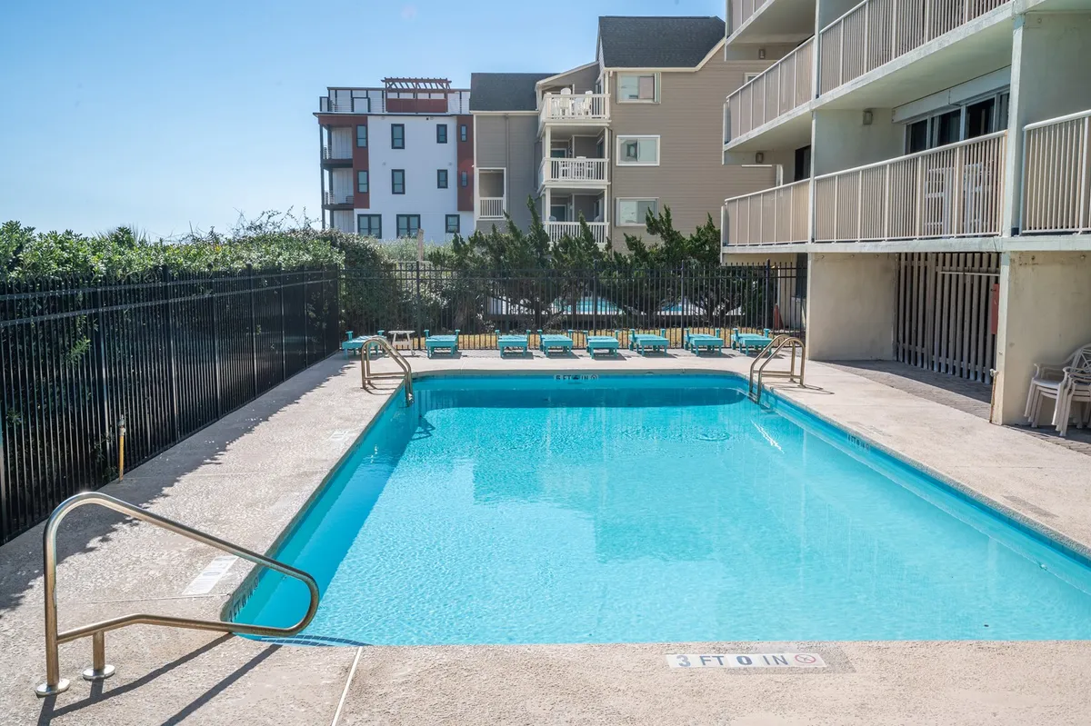An attractive outdoor pool scene framed by lush greenery, sun loungers, and apartment buildings. The pool is lively with people swimming and relaxing, evoking a leisurely atmosphere ideal for both active fun and quiet lounging.