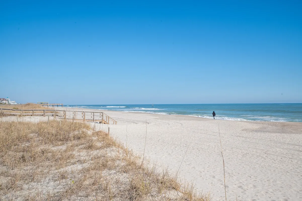 A serene exterior shot featuring sand dunes, rolling ocean waves, a wooden boardwalk, and a person walking along the shore. The scene captures the natural beauty and open space of the beach environment.