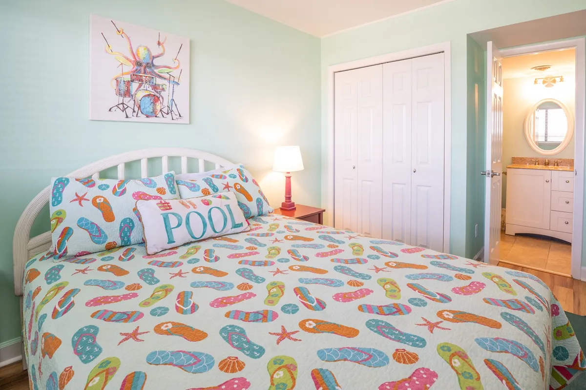 A playful bedroom featuring a twin bed with colorful flip-flop patterned bedding, cheerful wall art, and a bedside lamp. The space is bright and inviting, ideal for kids or singles seeking a cozy retreat.