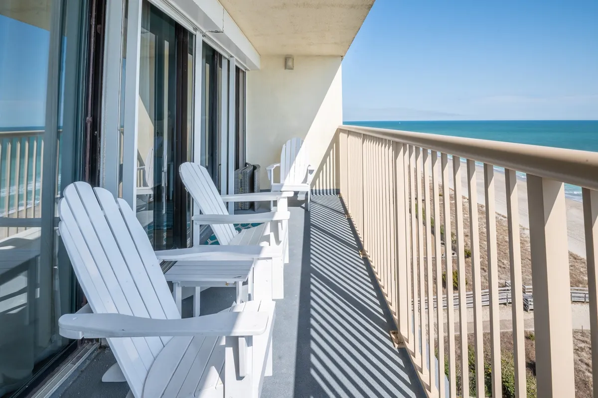 A serene balcony scene with white Adirondack chairs facing the ocean, bordered by a sturdy railing. The inviting setup is perfect for unwinding outdoors with views of the coast, embodying the relaxing, breezy evenings highlighted in the section.